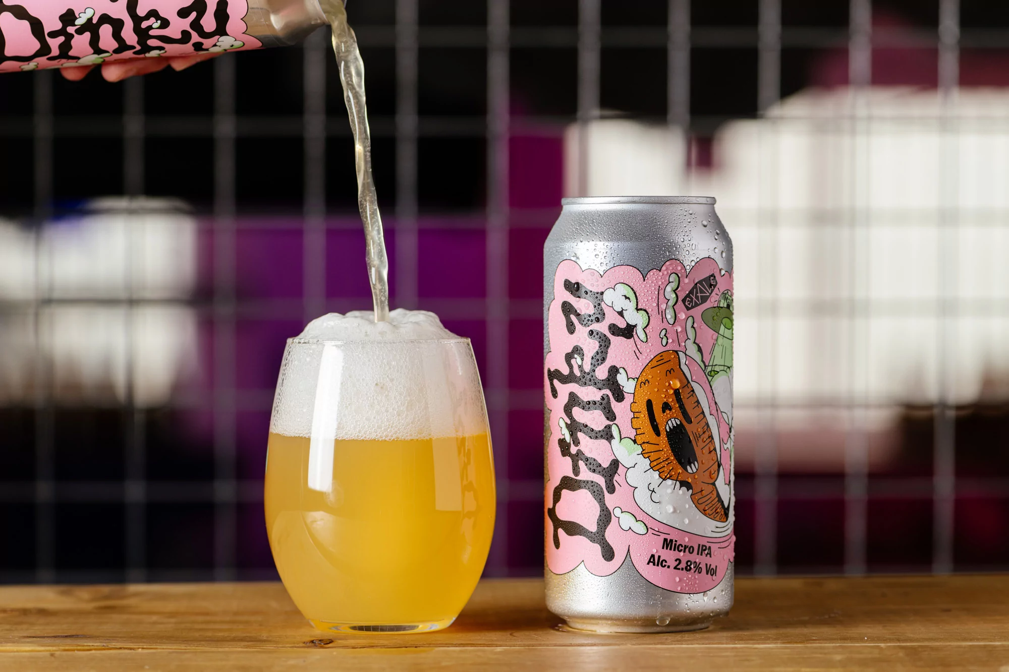 Beer is poured into glass beside a can of Dinky Micro IPA on a wooden surface against a black and purple background. Food and beverage.