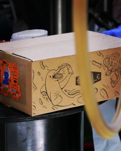 Cardboard box featuring cartoon drawings, including a shark holding a beverage and an alien, sits atop a dark, reflective surface.