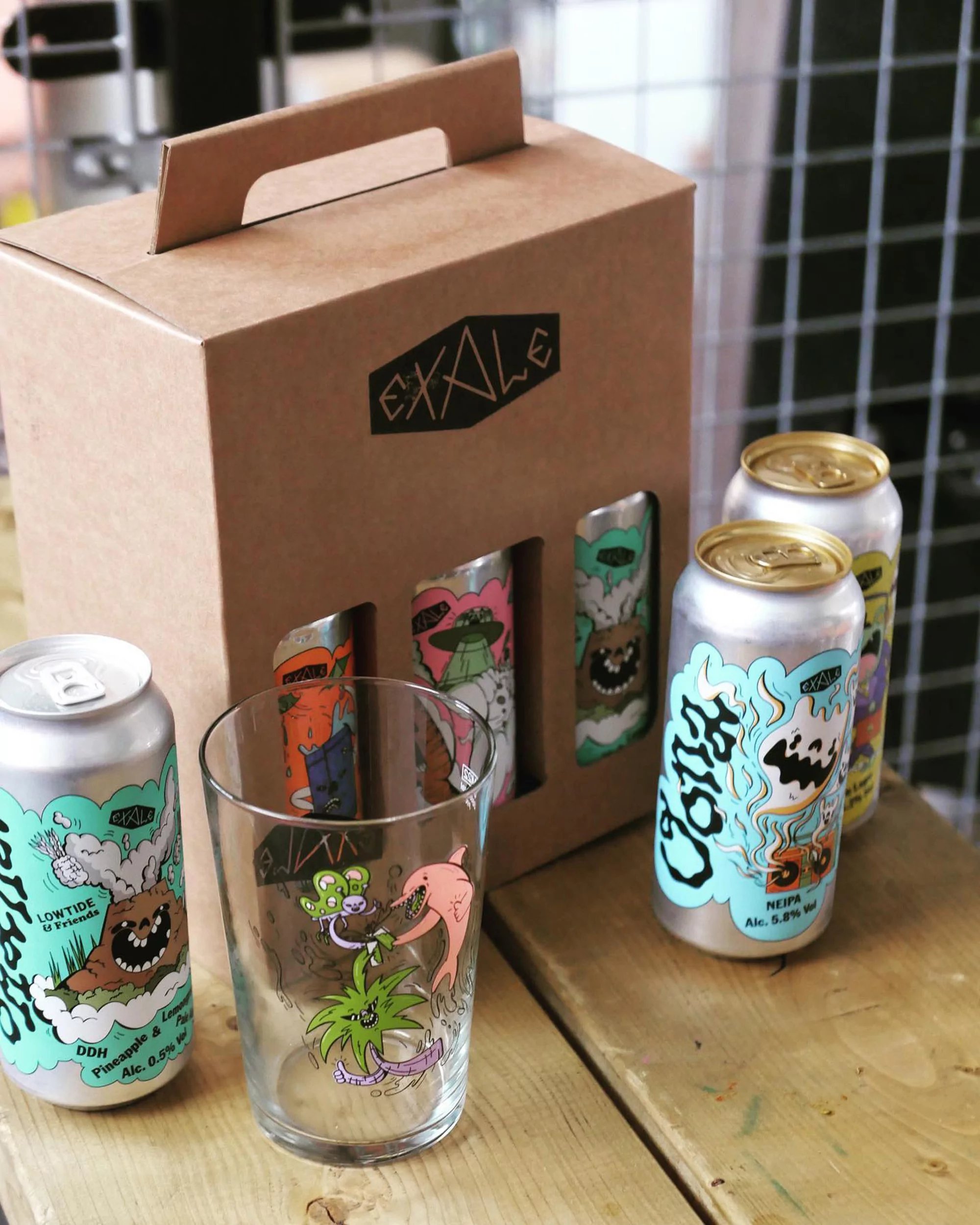 Several cans and a glass featuring monster illustrations on a wooden surface, near a cardboard box with 'EXALE' logo, indoor setting.