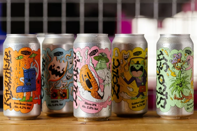 Five colorful beer cans with unique illustrated designs on a wooden surface in a studio setting, showcasing creative packaging for alcoholic beverages.