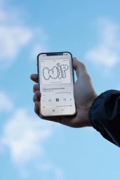 Hand holding a smartphone displaying an audio interface against a blue sky with soft clouds.