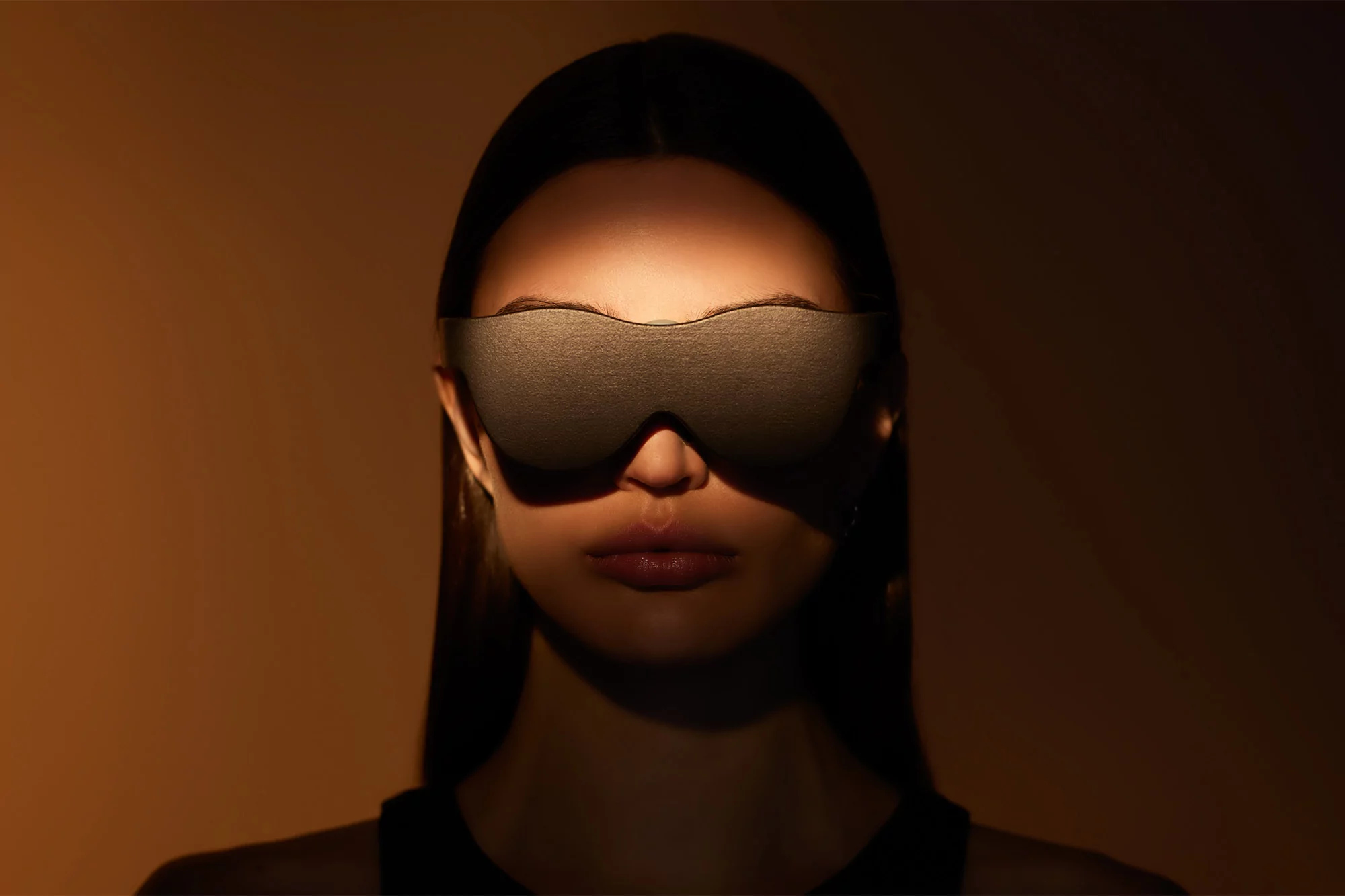 Close-up portrait of a woman wearing a black sleep mask in a dimly lit indoor setting with warm, golden tones.