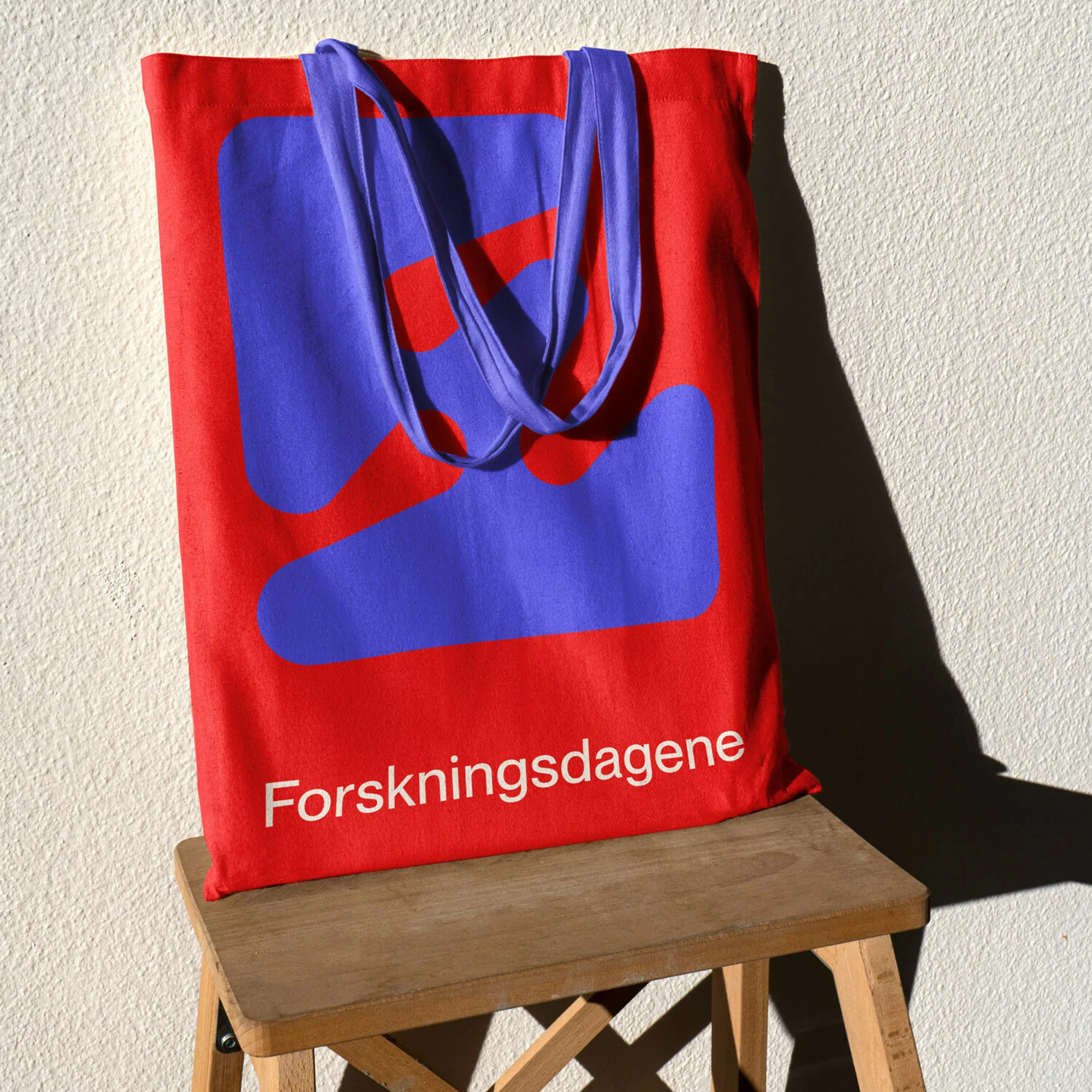 Red canvas tote bag with blue graphic and the text 'Forskningsdagene' on a wooden stool against a white wall, bright light.