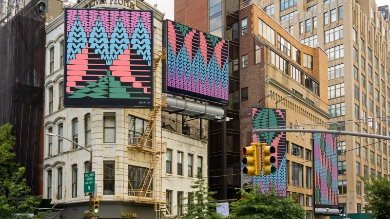 Outdoor view of buildings displaying graphic art billboards with abstract letters and shapes in pink, blue, and green in an urban environment.