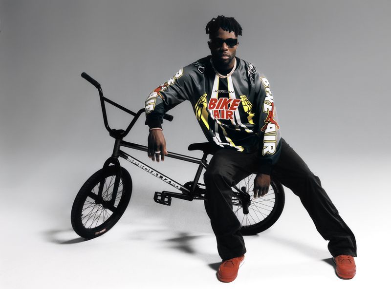 Man in racing jersey poses on black BMX bike in studio. He wears sunglasses and red sneakers in dynamic fashion shot.