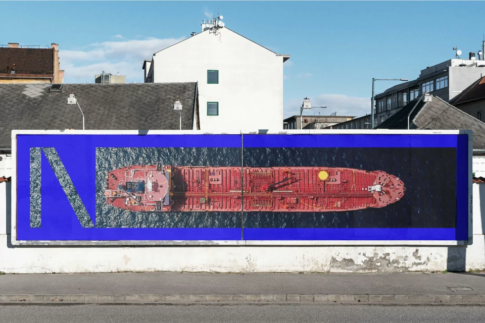 Aerial view of a red tanker ship is displayed on an outdoor billboard in an urban setting, under a clear blue sky.