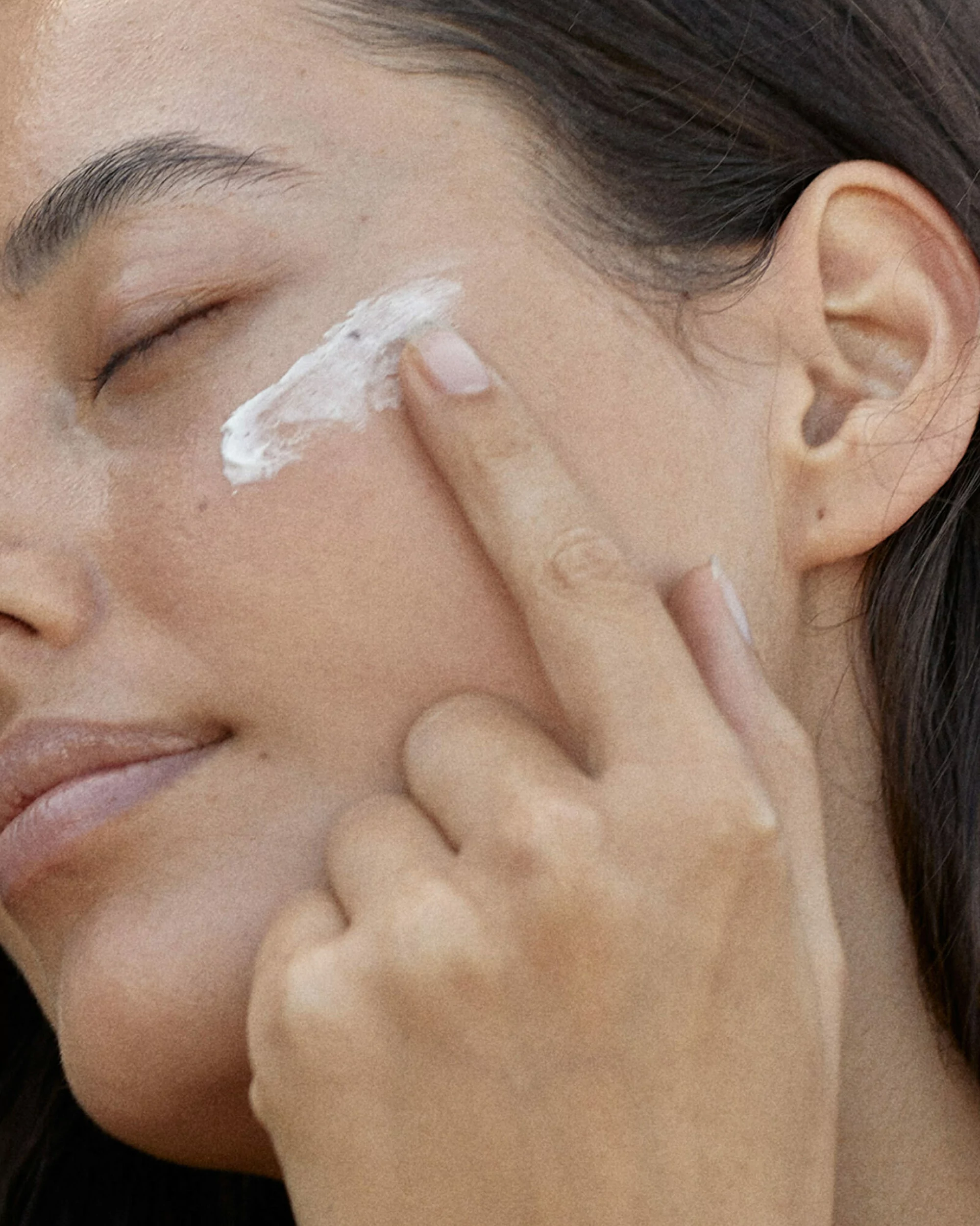 Close-up of a woman applying white cream to her face with her finger, emphasizing skincare and beauty, with focus on youthful complexion.