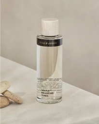 Clear skincare toner bottle with wooden cap standing upright on a pale textured surface beside three small stones