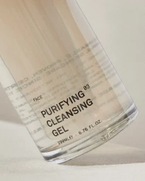 Close-up of a clear skincare bottle labeled purifying cleansing gel resting on a neutral textured surface