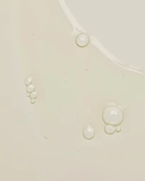 Close-up macro shot of transparent skincare serum liquid droplets resting on a smooth cream-colored surface.