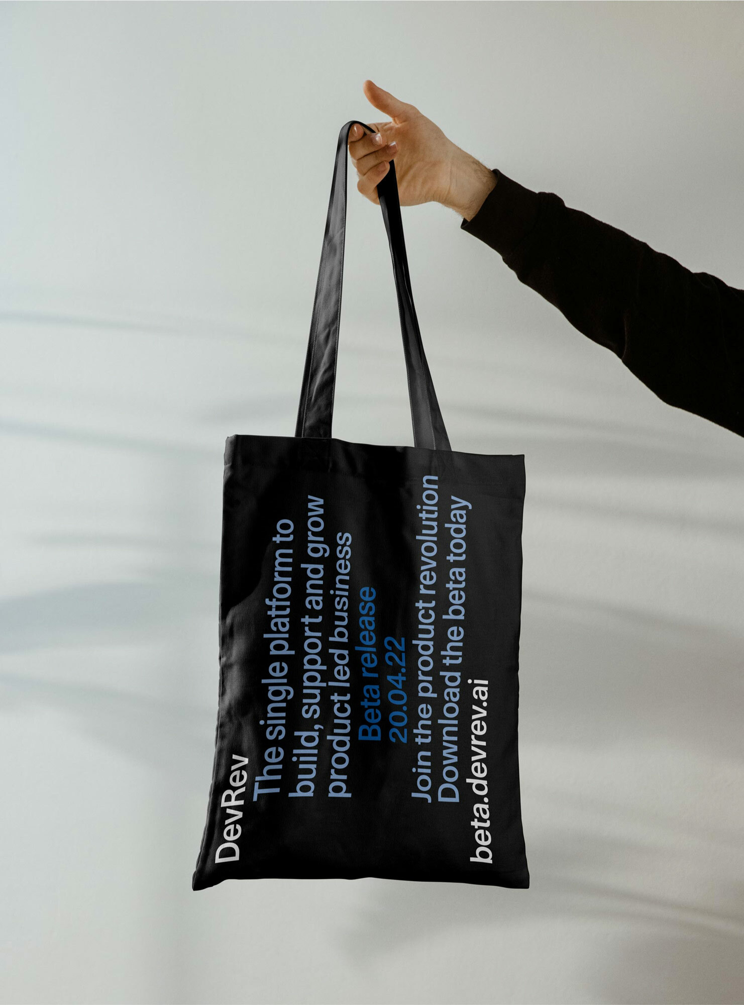 Hand holds a black tote bag with white text promoting DevRev, a platform for product-led business, against a light grey background.