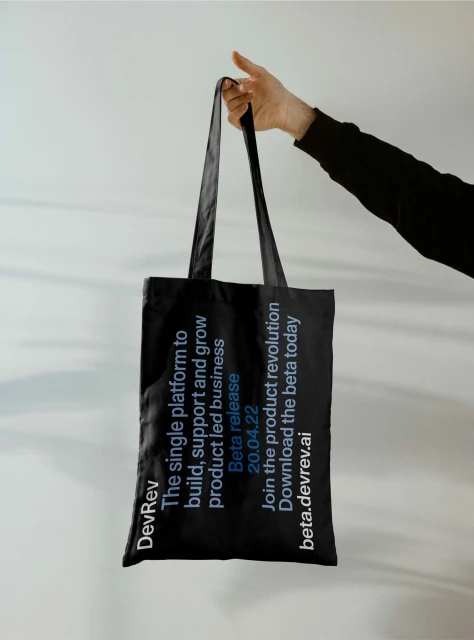 Hand holds a black tote bag with white text promoting DevRev, a platform for product-led business, against a light grey background.