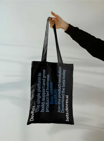 Hand holds a black tote bag with white text promoting DevRev, a platform for product-led business, against a light grey background.
