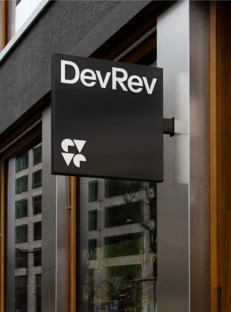 Black and white sign with 'DevRev' and logo outside a building, reflecting city buildings in the windows.