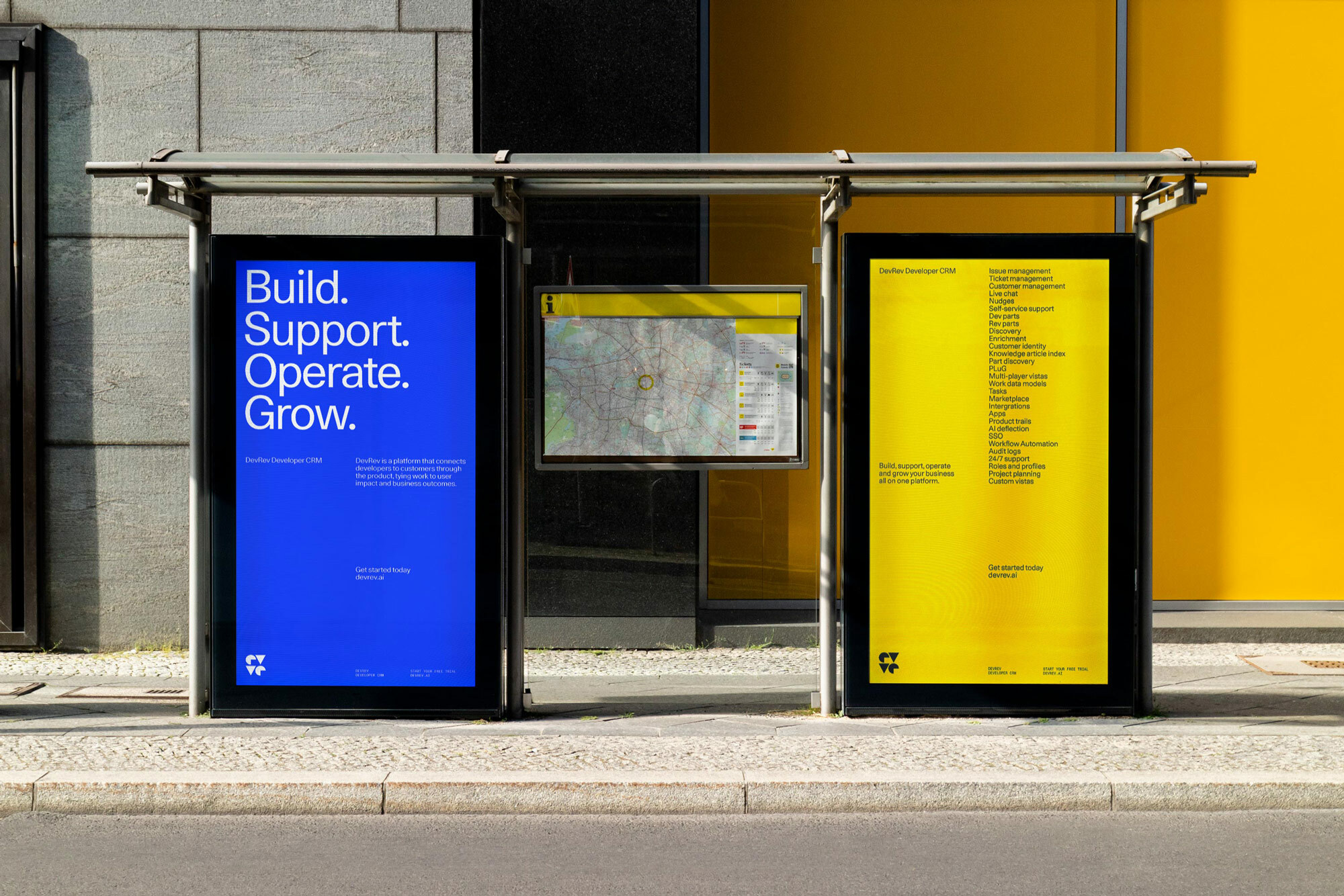 Bus stop advertisement displays blue and yellow posters with text and a map, next to a gray and yellow building facade. 
