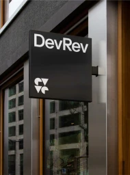 Black square storefront sign with white DevRev text mounted on a modern building exterior.