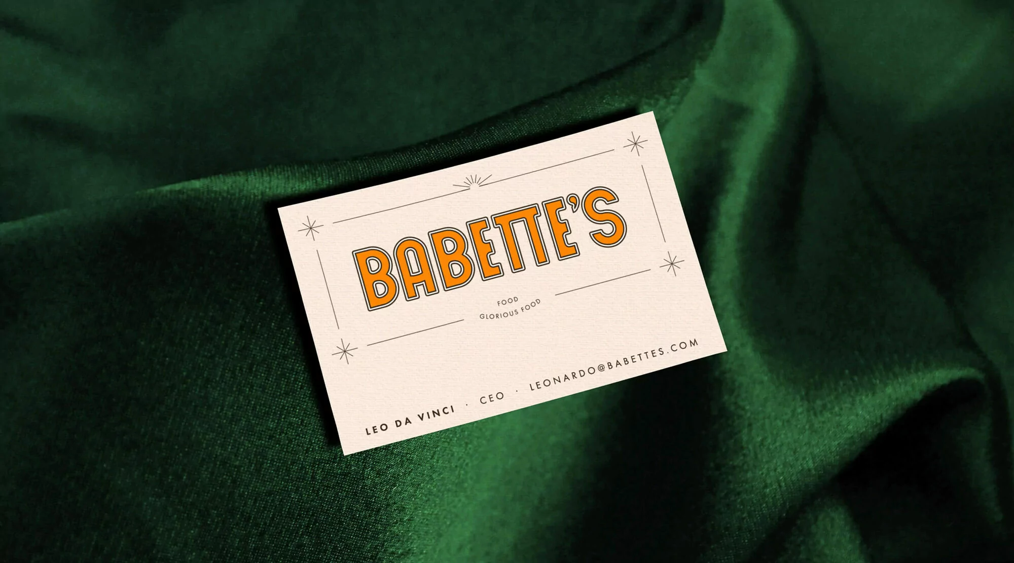 Business card featuring the logo “Babette’s FOOD GLORIOUS FOOD” in orange against a folded green velvet-like fabric background.