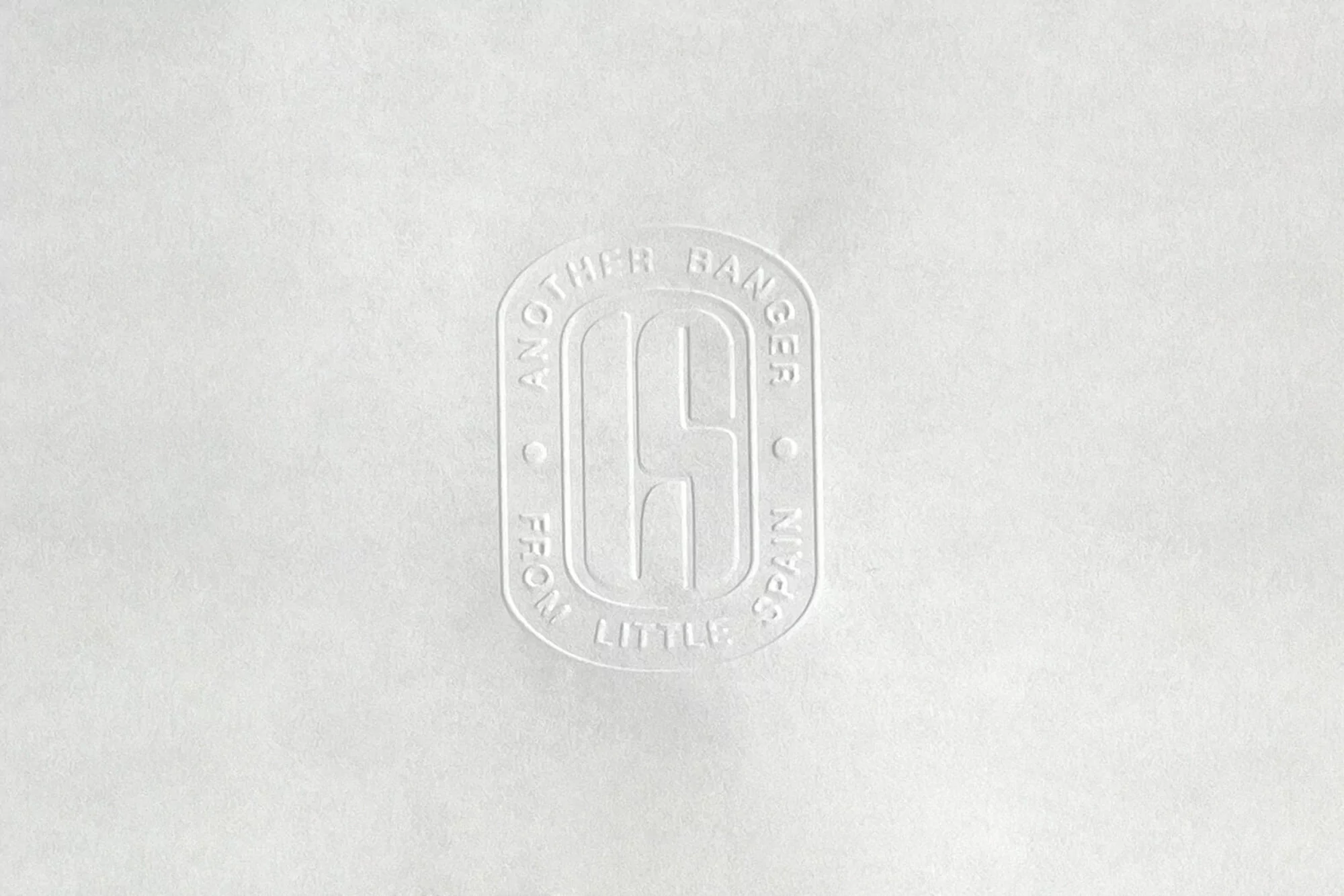 Close-up of embossed logo on white textured surface. The logo is oval-shaped and contains stylized text and a central 'S' symbol.