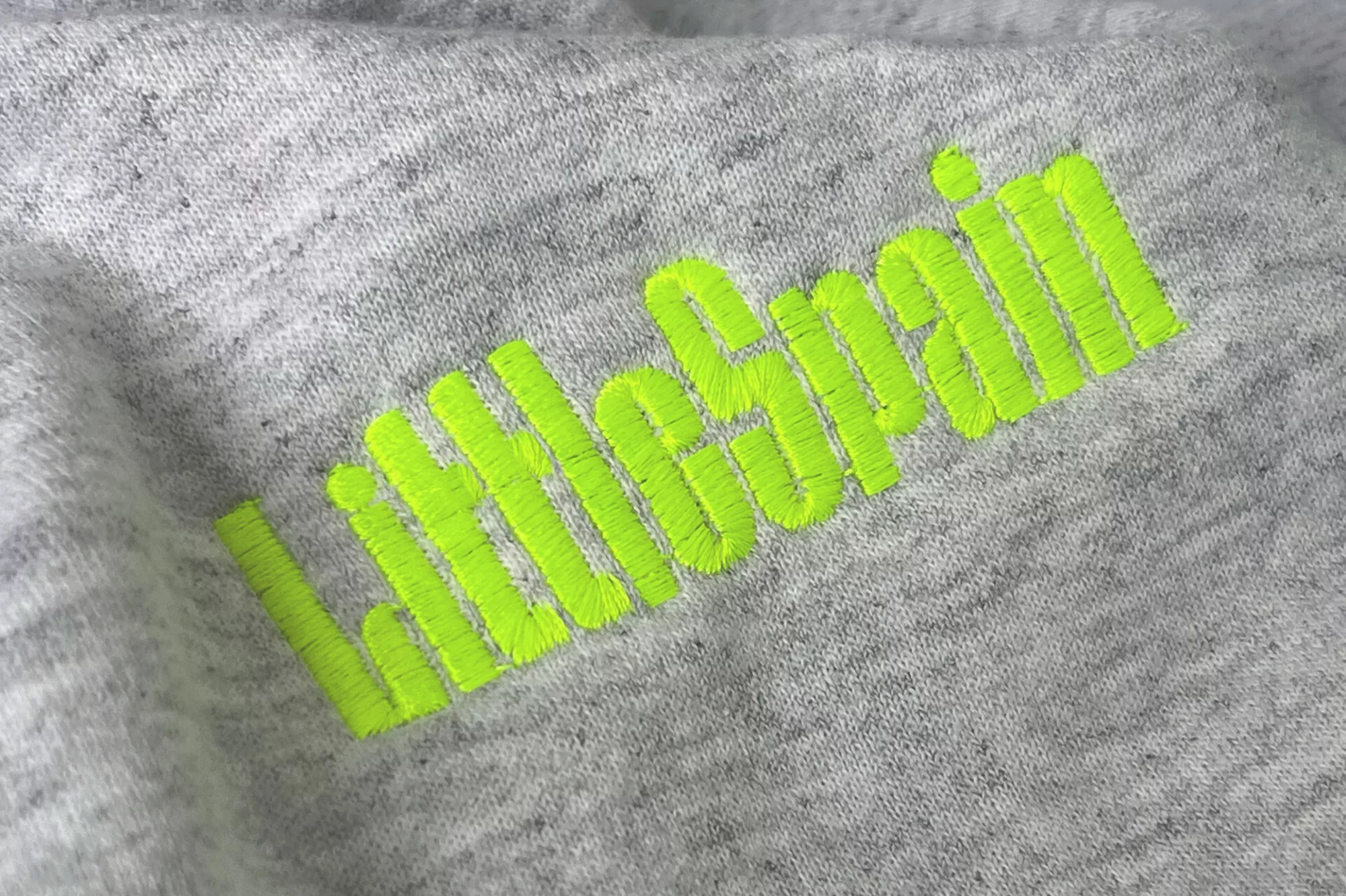 Close-up of a heather-grey heathered fabric with bright neon-green embroidered text that reads, 'Little Spain'.