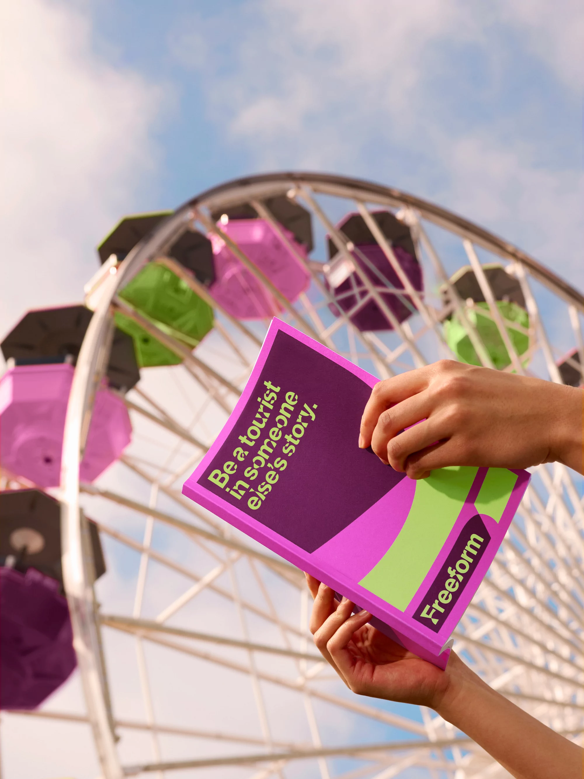 Hands holding a purple brochure with text 'Be a tourist in someone else’s story', featuring the Freeform logo against a Ferris wheel.