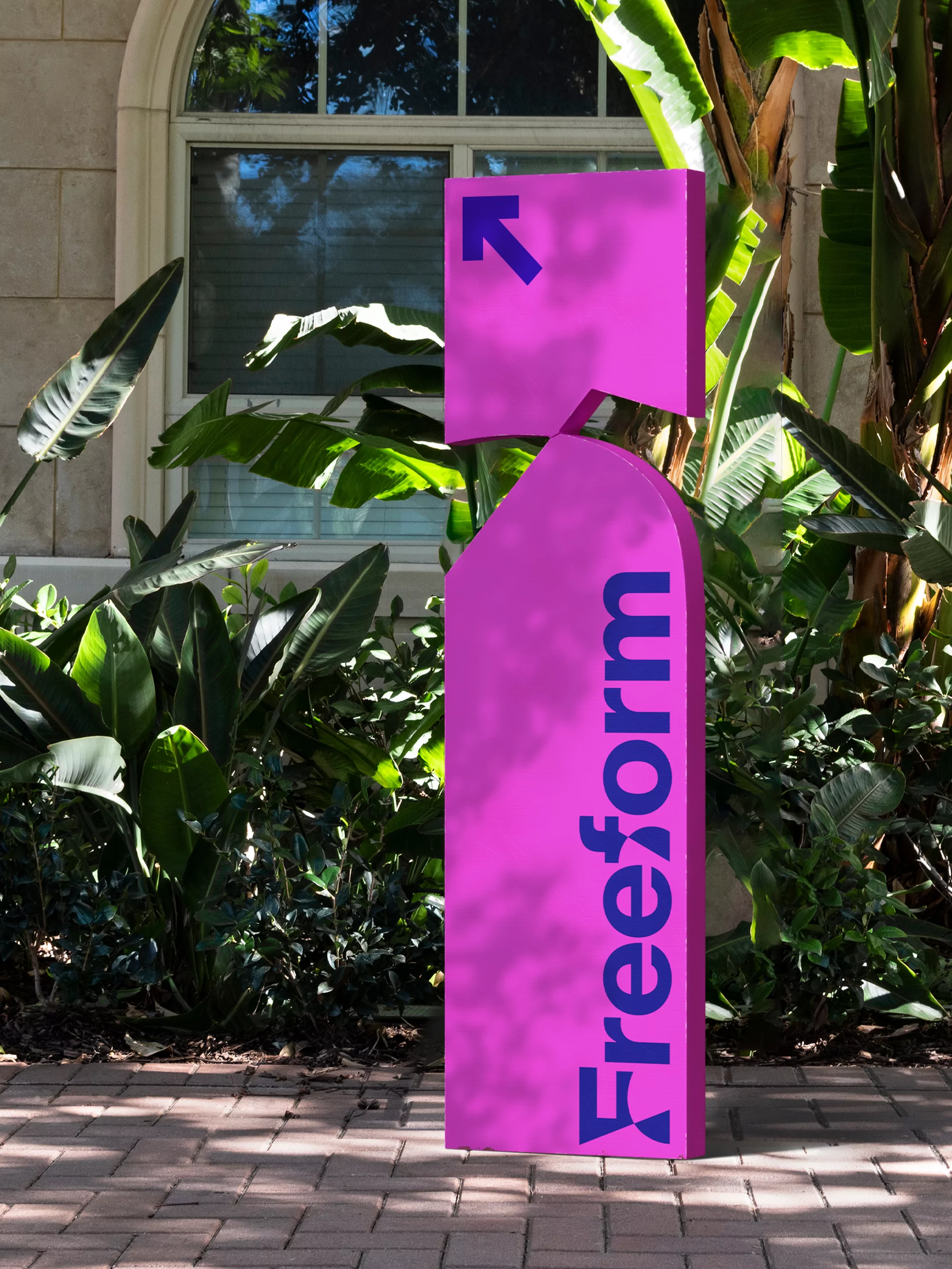 Outdoor shot of a purple sign that reads 'Freeform' in blue text, surrounded by lush green plants and trees on a sunny day.
