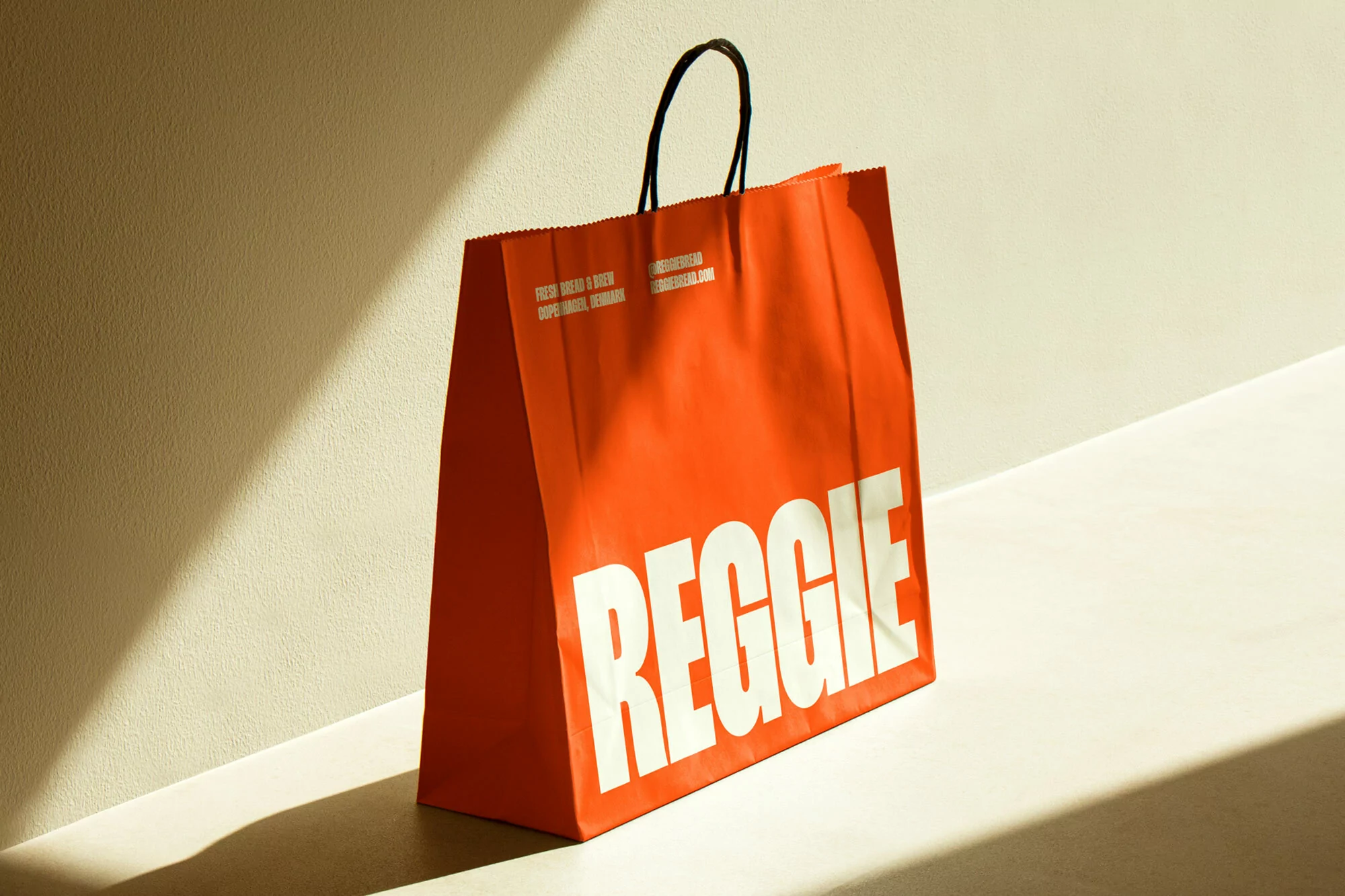 An orange shopping bag with the word 'REGGIE' in white letters sits in a sunlit indoor setting against a white wall and table.