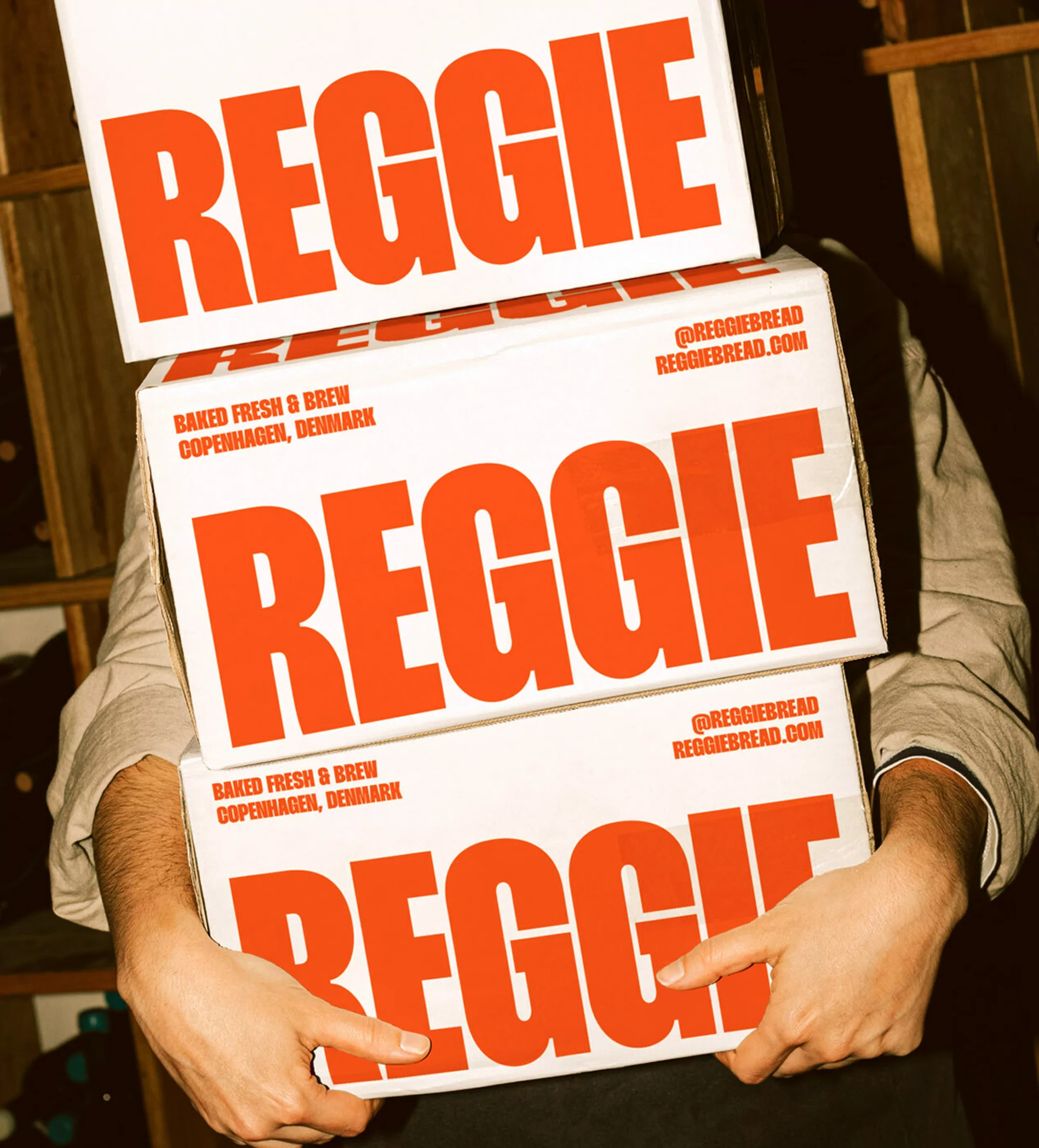 Person carrying three white boxes with 'REGGIE' in large orange letters. Text also says 'Baked Fresh & Brew, Copenhagen, Denmark'.