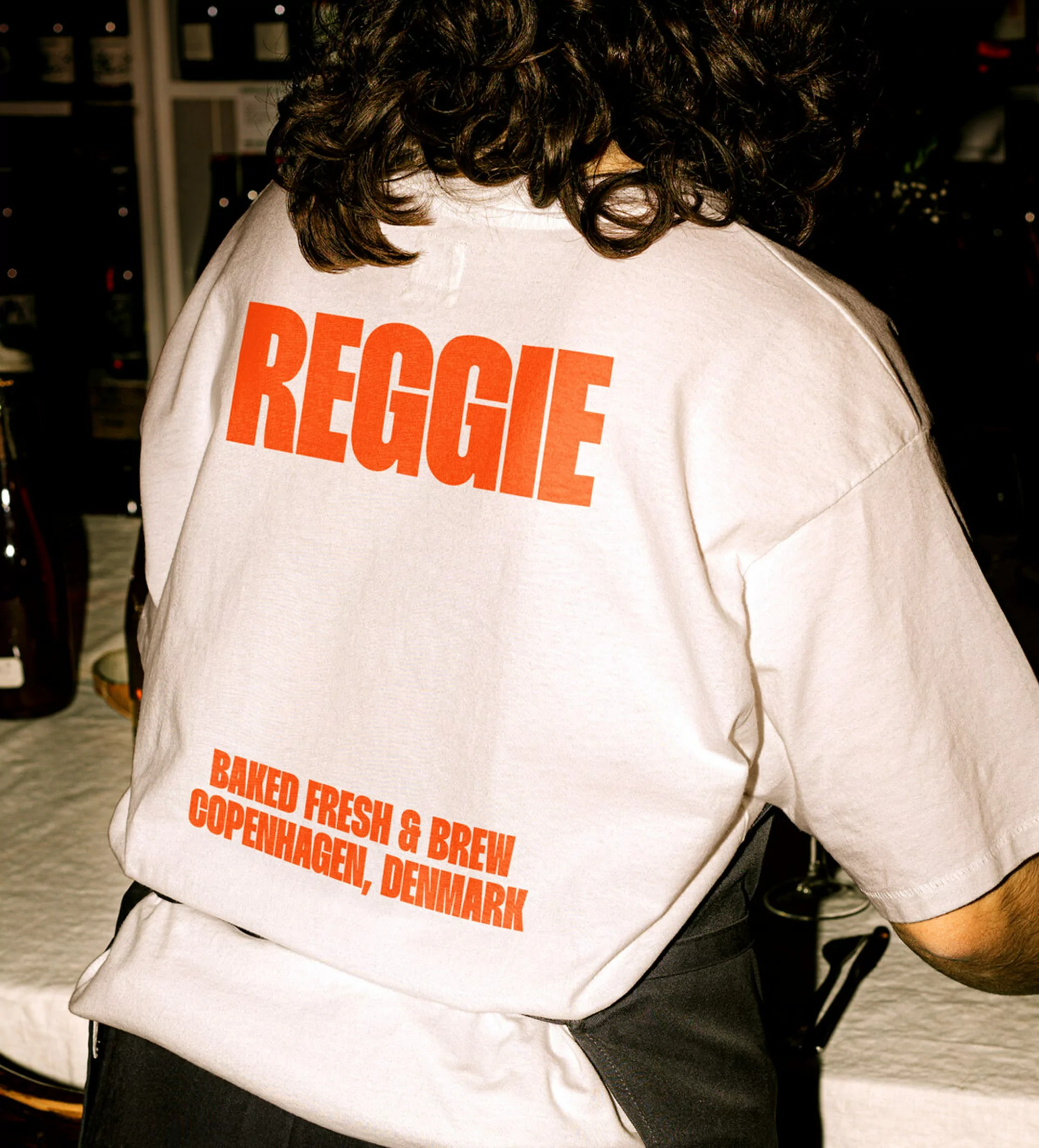Person seen from behind wearing a white t-shirt with orange REGGIE branding text against a softly lit indoor background.