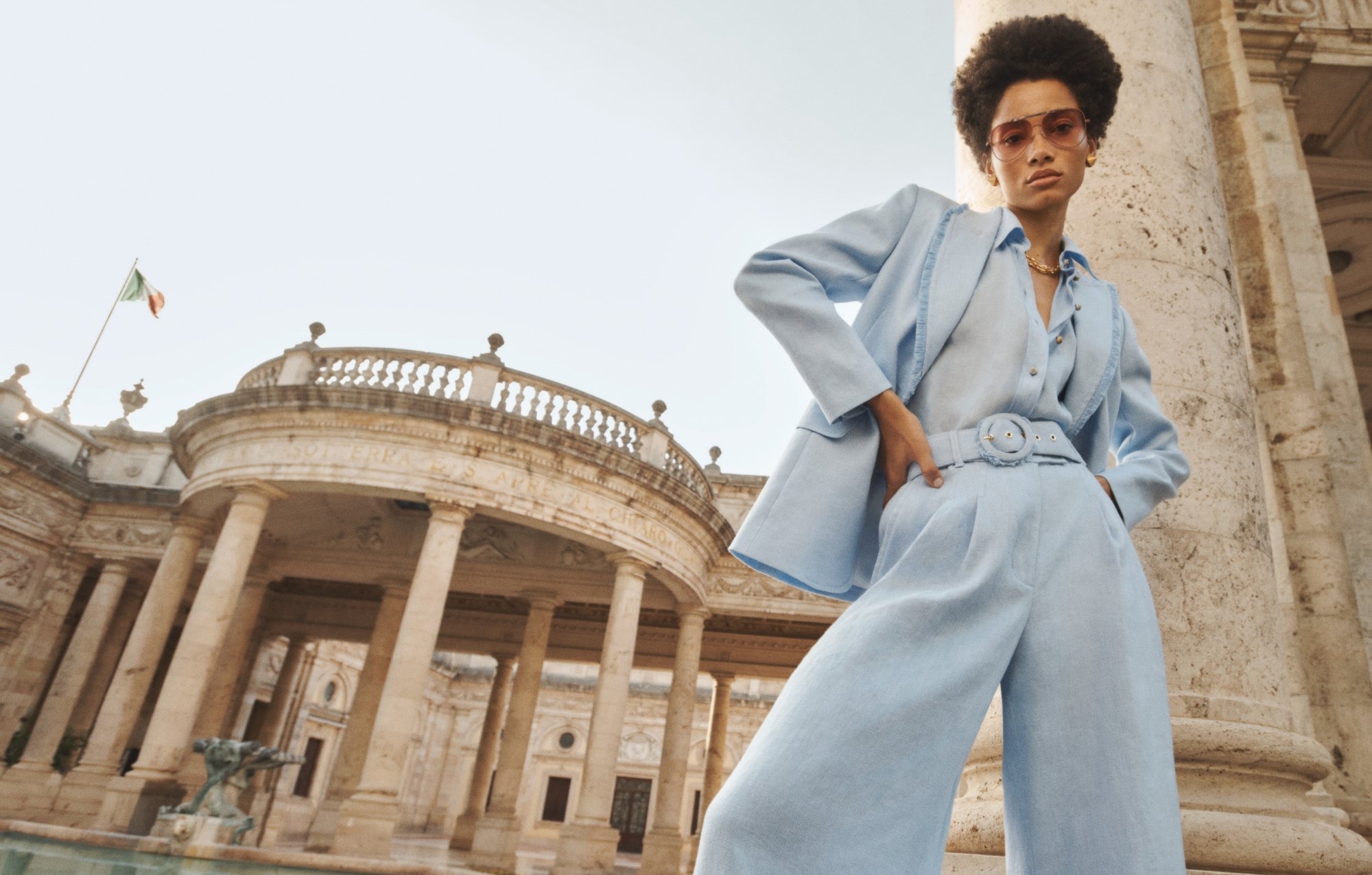 Fashion shot of an adult woman in a light blue suit standing outside with a historic building and Italian flag in the background.