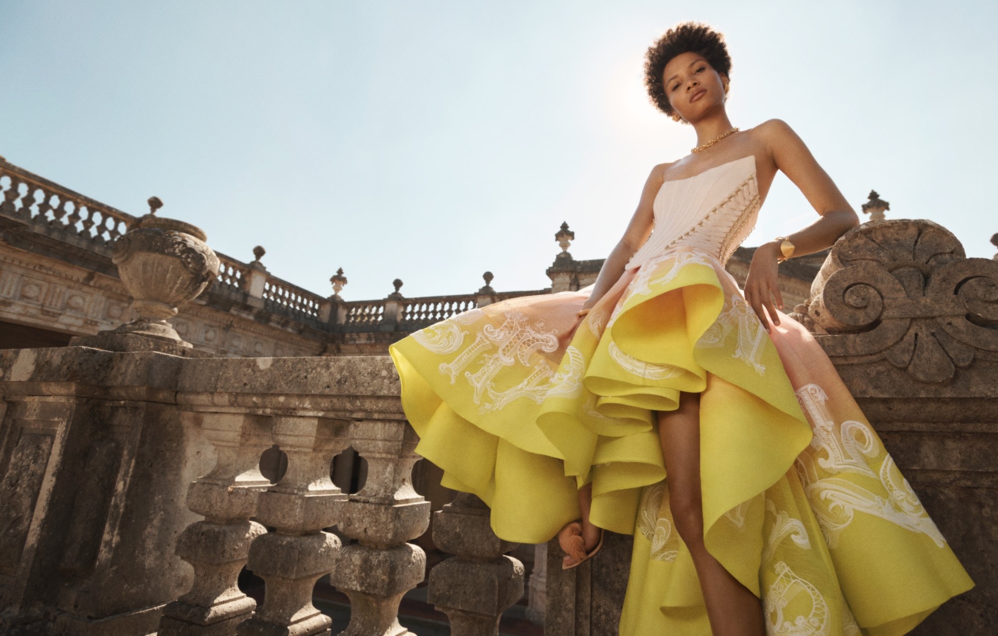 Woman posing in an elaborate yellow gown with white details on a stone terrace with balustrades under a bright blue sky.