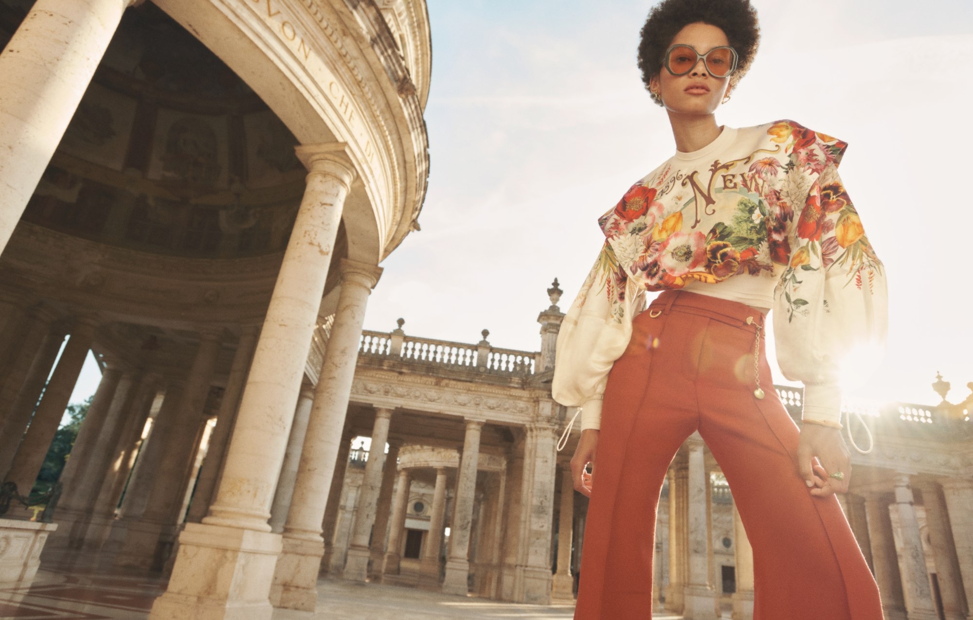 A woman with sunglasses models floral top and terracotta pants near a colonnade under a bright sky, fashion editorial, travel photography.
