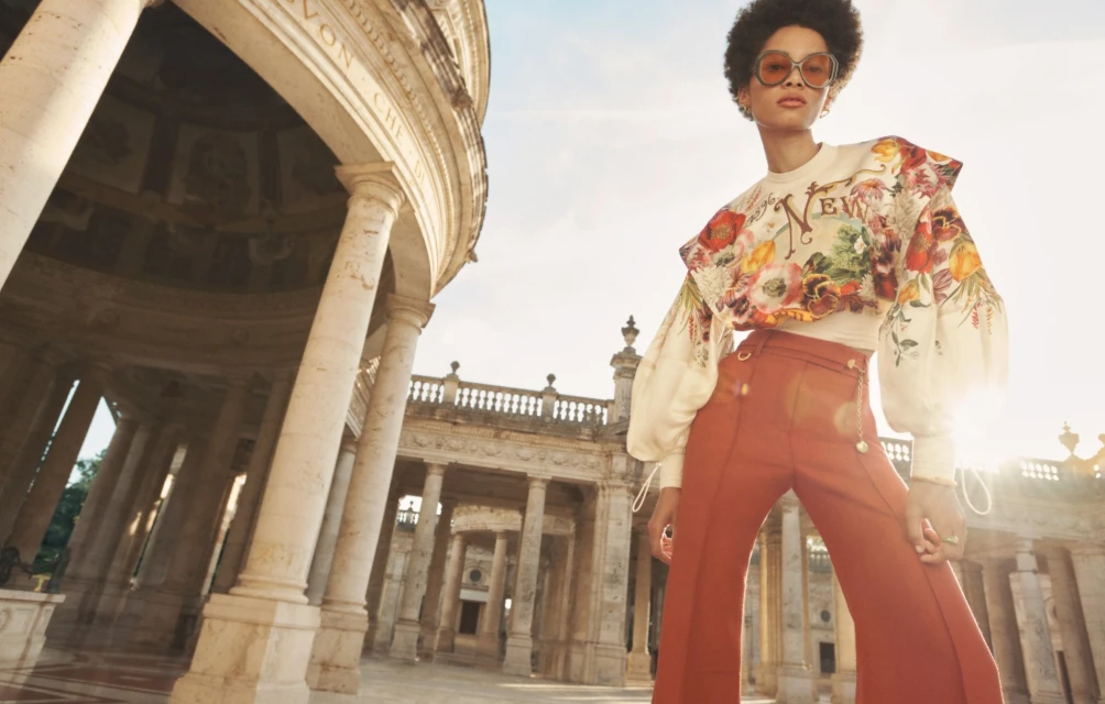 A woman with sunglasses models floral top and terracotta pants near a colonnade under a bright sky, fashion editorial, travel photography.