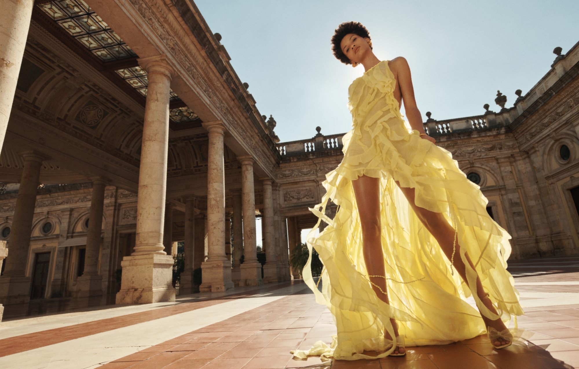 Fashion shot of an adult female wearing a yellow gown with ruffles, standing in front of colonnades with an ornate background.
