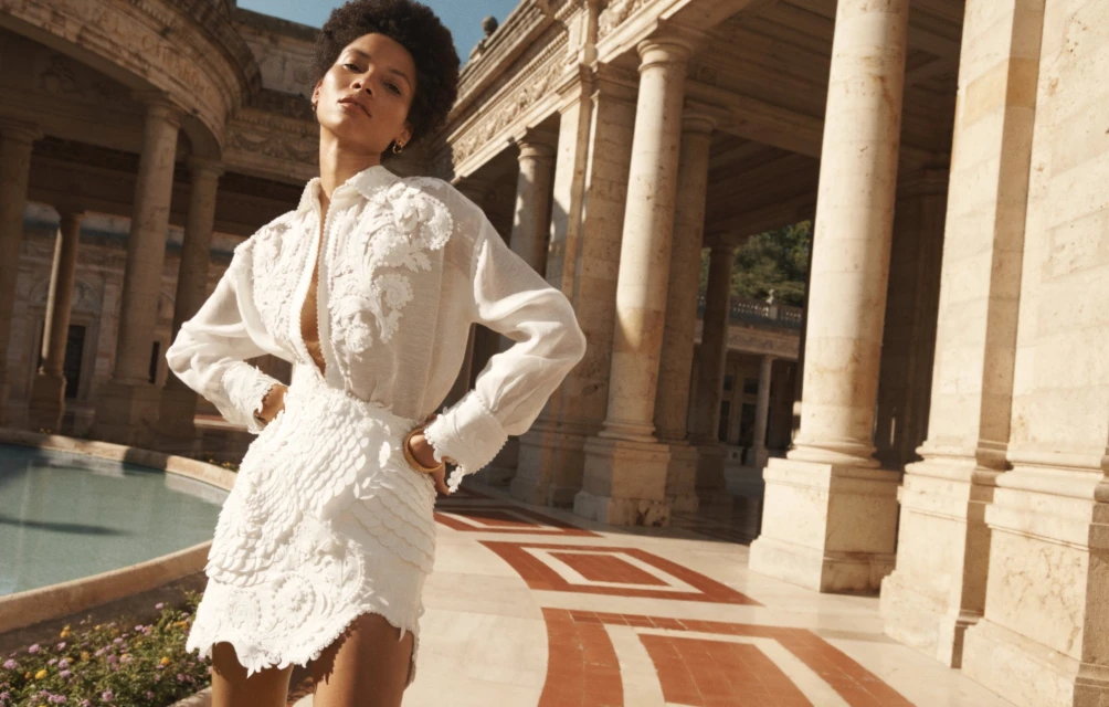A stylish woman in a white linen dress stands in a colonnaded courtyard with geometric paving, bathed in warm, natural light.