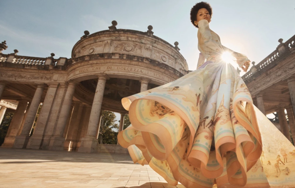 Woman in a flowing gown posing near an ancient Roman building on a sunny day. Fashion shoot against an architectural backdrop.