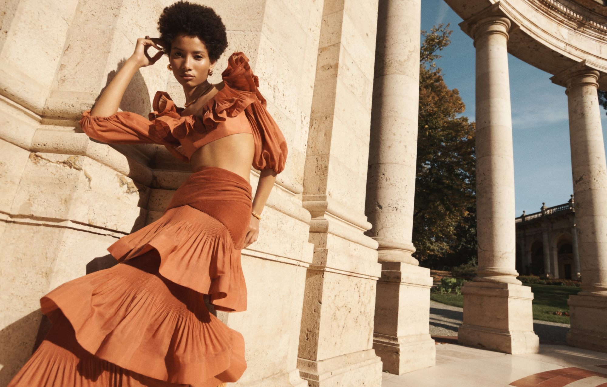 Elegant African-American woman in a rust-colored two-piece outfit poses against the stone columns of an outdoor arcade on a sunny day.