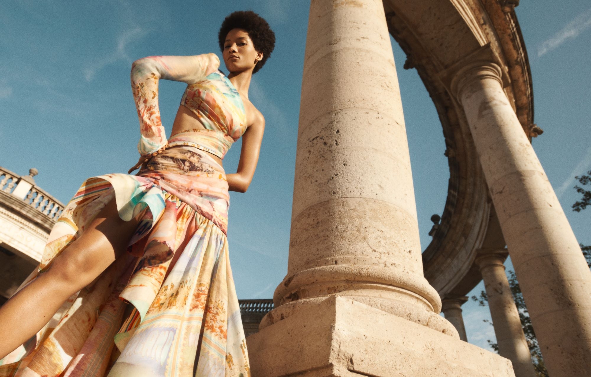 A woman in a colorful dress poses next to stone columns under a blue sky, showcasing fashion in an outdoor architectural setting.