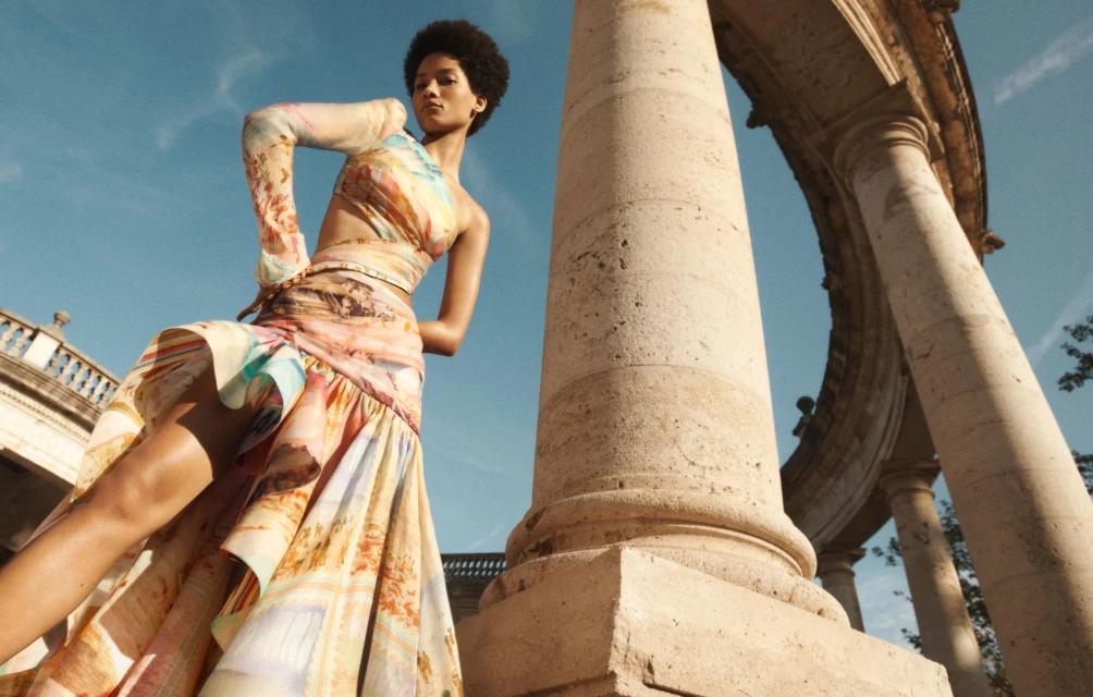 A woman in a colorful dress poses next to stone columns under a blue sky, showcasing fashion in an outdoor architectural setting.