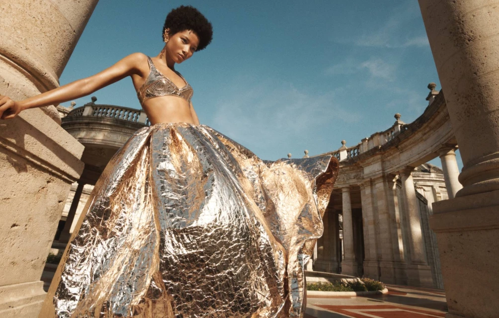 An adult female model poses in a metallic silver and gold gown, standing outdoors against a backdrop of an old stone building with columns.