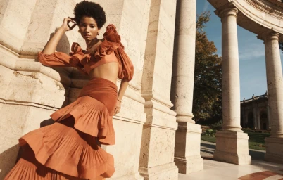 Model Lineisy Montero in a rust-colored ruffled crop top and tiered maxi skirt leaning against stone architectural columns.