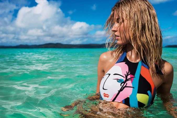Woman in swimwear with wet blonde hair partially submerged in ocean with blue sky. The swimsuit has an abstract face design on it.