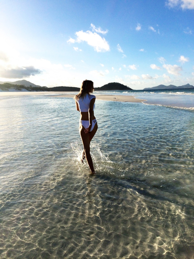 Model Martha Hunt in a white bikini walking through shallow turquoise water on a sandy beach under a blue sky