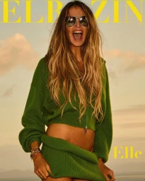 A woman wearing a green sweater and sunglasses laughing outdoors under a sunset sky for Elbazin Magazine.