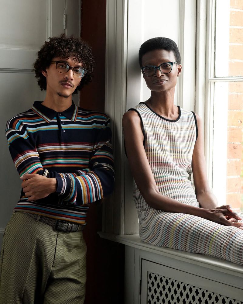 Two models pose indoors by a window in striped knitwear and eyeglasses, radiating a fashion-forward, sophisticated, and eye-catching aesthetic.