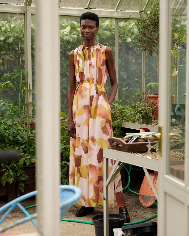 Woman in a printed dress stands inside a glass-walled greenhouse surrounded by plants, garden tools, and natural light.