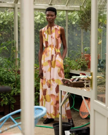 Woman in a printed dress stands inside a glass-walled greenhouse surrounded by plants, garden tools, and natural light.