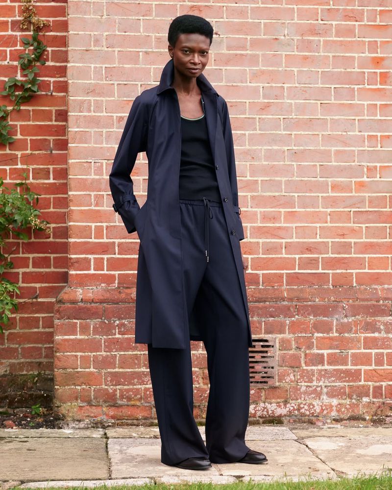 An adult female model poses in a long navy trench coat and matching pants against a brick wall with ivy in a medium outdoor shot.