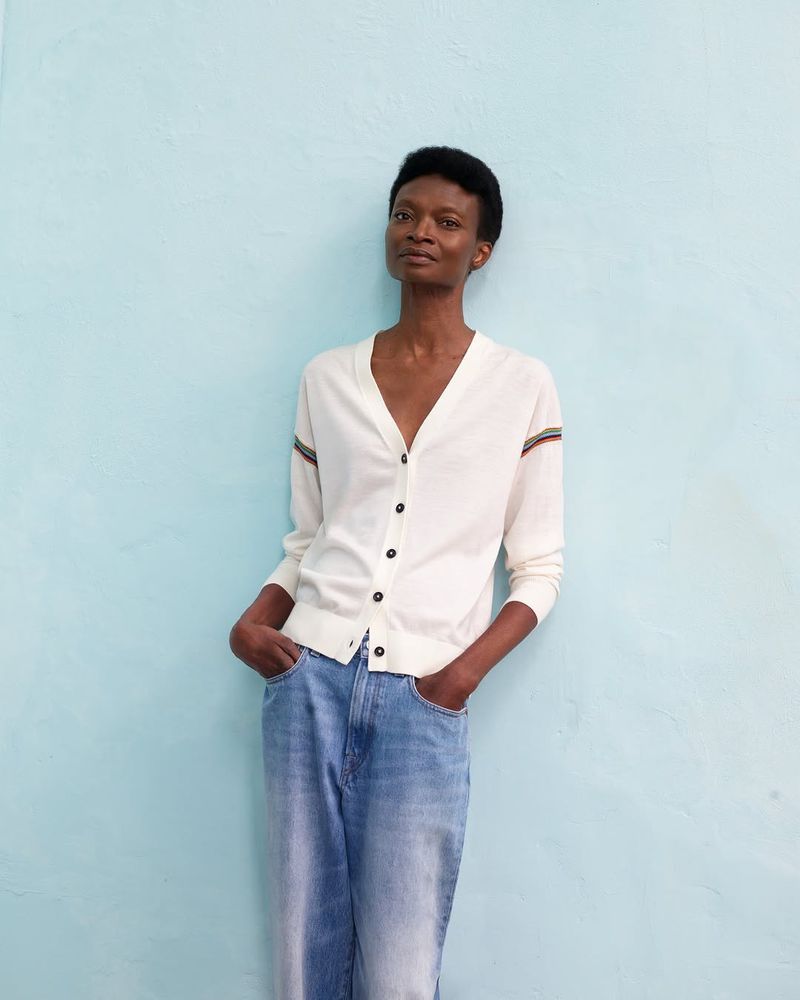 Woman in a cream-colored cardigan and jeans stands against a blue wall. Fashion portrait with casual style and solid background.