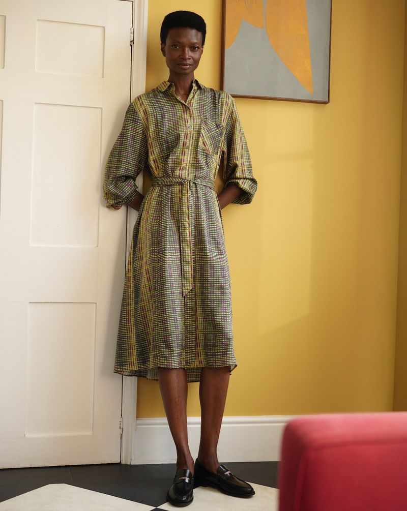 Elegant Black woman poses in yellow plaid dress with belt and black loafers against yellow wall with abstract art and white door.