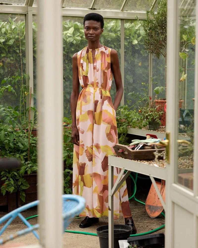 Model Debra Shaw wearing a floral print sleeveless dress standing inside a greenhouse for Paul Smith